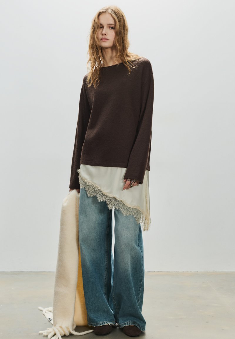 Barna hosszú ujjú felső, krémszínű, aszimmetrikus, csipkés szegélyű inggel rétegezve. Széles szárú kék farmer és semleges színű sál egészíti ki a ruhát.