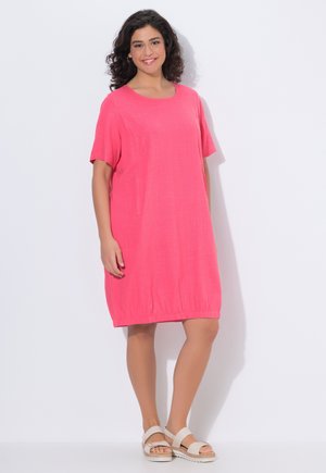 Donna sorridente, con un vestito rosa largo fino al ginocchio e sandali bianchi, in piedi su uno sfondo bianco.