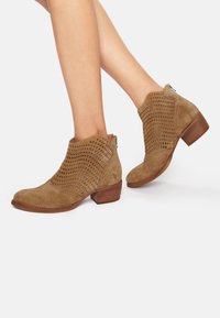 Bottines en suède beige avec perforations découpées, bout rond et petit talon empilé, dotées d'une fermeture éclair arrière pour un enfilage facile.