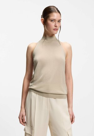 Femme aux cheveux lissés en arrière, portant un haut beige sans manches à col montant et un pantalon large crème sur un fond blanc uni.