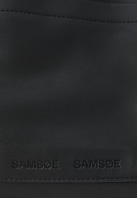 Materiale in pelle nera con una superficie testurizzata. Logo "SAMSOE SAMSOE" impresso in un colore scuro, design minimalista con dettagli di cucitura discreti.