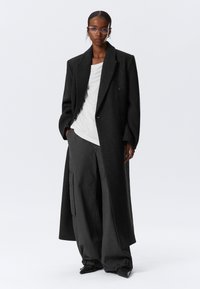 Langer schwarzer Wollmantel, doppelt knöpfbares Design, strukturiertes Material. Darunter ein weißes Top und eine lockere graue Hose mit Taschen.
