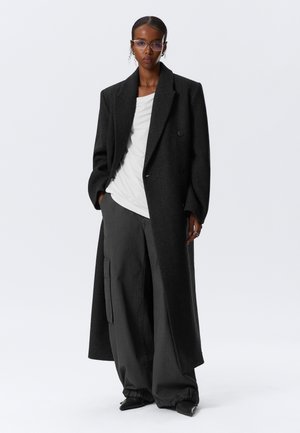 Manteau long en laine noire avec un design croisé, associé à un haut blanc et un pantalon large gris foncé. Présente des chaussures pointues élégantes et des lunettes.