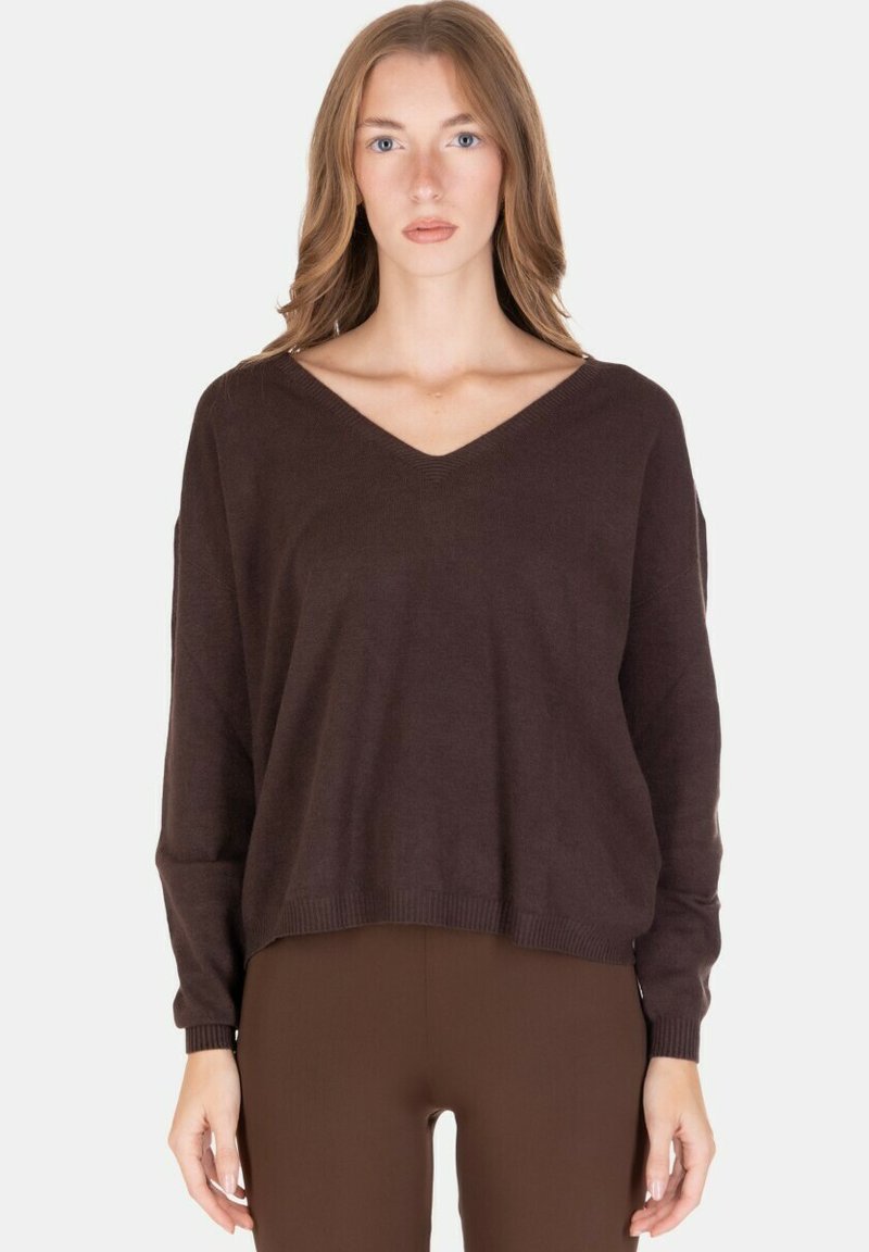 Donna che indossa un maglione a collo a V ampio di colore marrone scuro e pantaloni marroni abbinati, in piedi davanti a uno sfondo chiaro e uniforme.