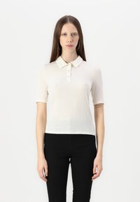 Kortermet polo skjorte i krem, med knappelukning, myk tekstur og en figurnær design, brukt sammen med svarte bukser.