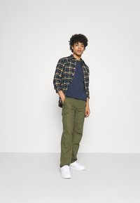 Camisa de flanela xadrez em azul-marinho, verde e amarelo, sobre uma t-shirt azul-marinho, combinada com calças cargo em verde-azeitona e ténis brancos.