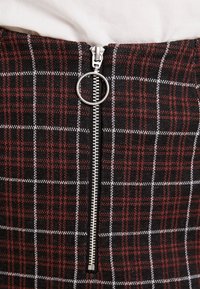 Metalen rits met ronde trekker op rood en zwart geruite stof, gedragen met een witte top.