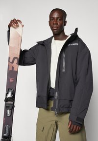 Giacca da sci nera impermeabile con cappuccio, zip anteriore e dettaglio del logo. Uomo che tiene uno sci marrone e nero con attacchi. Pantaloni verde oliva.