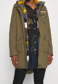Parka verde oliva con capucha de piel sintética, cierre de cremallera y múltiples bolsillos; sobre una camisa abotonada de flores oscuras con acentos azules.