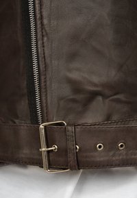 Dettaglio di una giacca di pelle marrone con una cerniera argentata, una cintura con una fibbia in metallo e una texture liscia con minime pieghe.