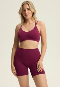 Burgundowy sportowy biustonosz z prążkowanego materiału z regulowanymi ramiączkami w komplecie z dopasowanymi szortami o wysokim stanie, charakteryzującymi się bezszwowym wzorem i miękką teksturą.