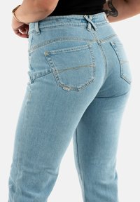Vaqueros de mezclilla azul claro con una cintura alta, bolsillos traseros prominentes y sutiles detalles de costura amarilla. Presentan un corte relajado y una textura suave.