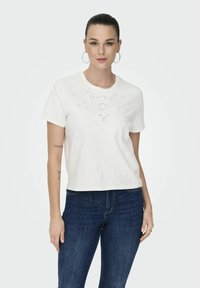 Hvid bomulds t-shirt med korte ærmer, med udskårede blomster mønstre på forsiden og en afslappet pasform, parret med mørkeblå jeans.