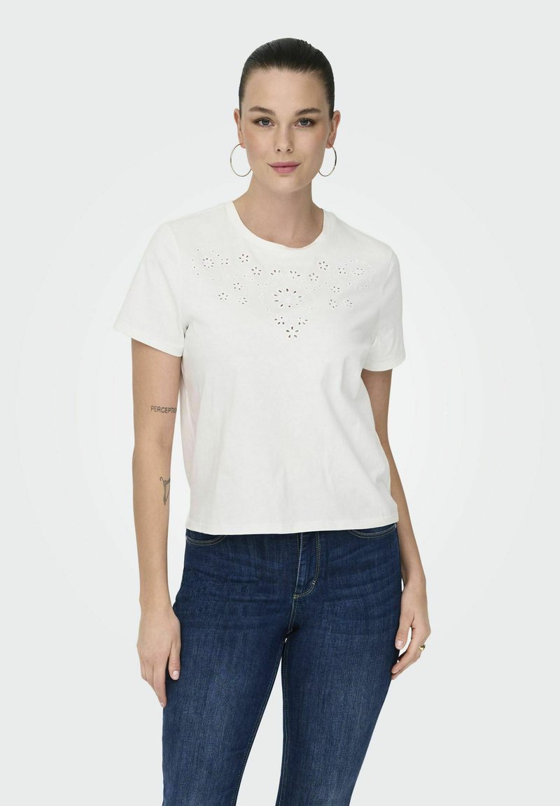 Hvid bomulds t-shirt med korte ærmer, med udskårede blomster mønstre på forsiden og en afslappet pasform, parret med mørkeblå jeans.