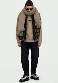 Suéter beige bajo un chaleco negro con estampado, bufanda gris, pantalones negros y zapatos beige. Los accesorios incluyen gafas de sol.