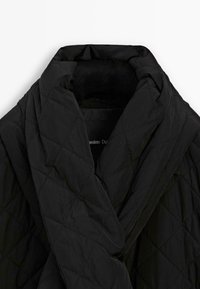 Svart quiltad jacka med stor, vadderad sjalkrage och diamantbroderimönster, vikta i en mjuk lagerstil.