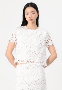 Top corto in pizzo bianco con motivi floreali ritagliati, maniche corte e scollatura tonda, abbinato a una gonna in pizzo coordinata.