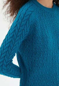 Turkos stickad tröja med texturerade mönster, som har ett kabelmönster på ärmarna och en rund halsringning.