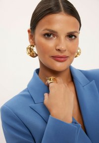 Złote kolczyki w formie statement, złoty pierścionek i niebieska marynarka. Kolczyki mają organiczny kształt; marynarka charakteryzuje się strukturalnym fasonem i klapami.