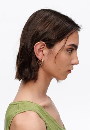 Kobieta z krótkimi brązowymi włosami, ubrana w zielony top bez rękawów i złotą skręconą kolczyk kołowy w lewym uchu, widziana z profilu na białym tle.