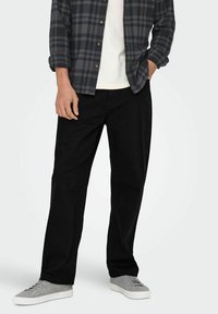 Svarta byxor med rak linje, kombinerat med en mörk rutig flanellskjorta och grå skor. Outfiten präglas av en avslappnad stil.