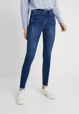 Femme portant un jean skinny bleu foncé avec une légère usure, des baskets blanches et une chemise à rayures bleues à manches longues.