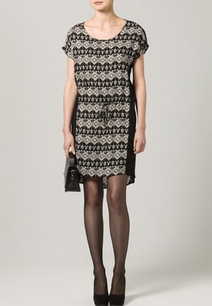 Robe à motifs noirs et crème avec des manches courtes, taille cintrée et panneaux latéraux noirs unis ; associée à des chaussures à talons noires et un sac à main.