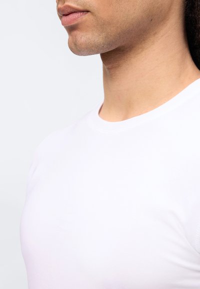 Πλευρική όψη του κάτω μέρους του προσώπου, του λαιμού και του άνω μέρους του στήθους ενός ατόμου που φοράει απλό λευκό μπλουζάκι με στρογγυλή λαιμόκοψη, σε απλό φόντο.