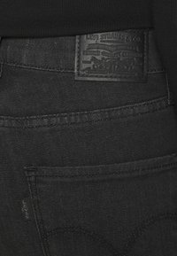 Vaqueros de mezclilla negra con una superficie texturizada, costuras visibles y una etiqueta de parche de cuero con detalle en relieve en la cinturilla.