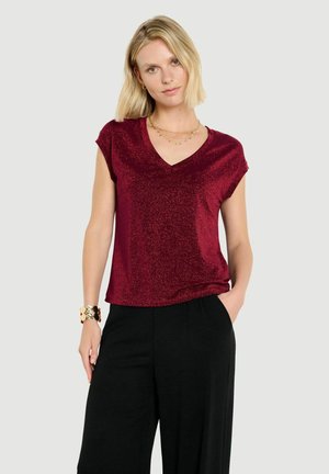 Femme aux cheveux blonds courts portant un haut bordeaux brillant à manches courtes et col en V, ainsi qu’un pantalon noir, debout, une main dans la poche.