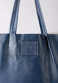 Blue leather tote bag with smooth texture, featuring two sturdy straps and a rectangular label with four metallic studs.