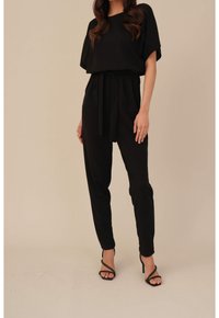 Femme portant une combinaison noire ceinturée à manches courtes et des sandales à talons hauts noires à lanières sur un fond neutre.