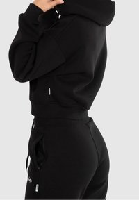 Felpa nera a vestibilità ampia, con cappuccio regolabile e polsini a coste. Abbinata a pantaloni jogger neri coordinati, con tasche con zip.