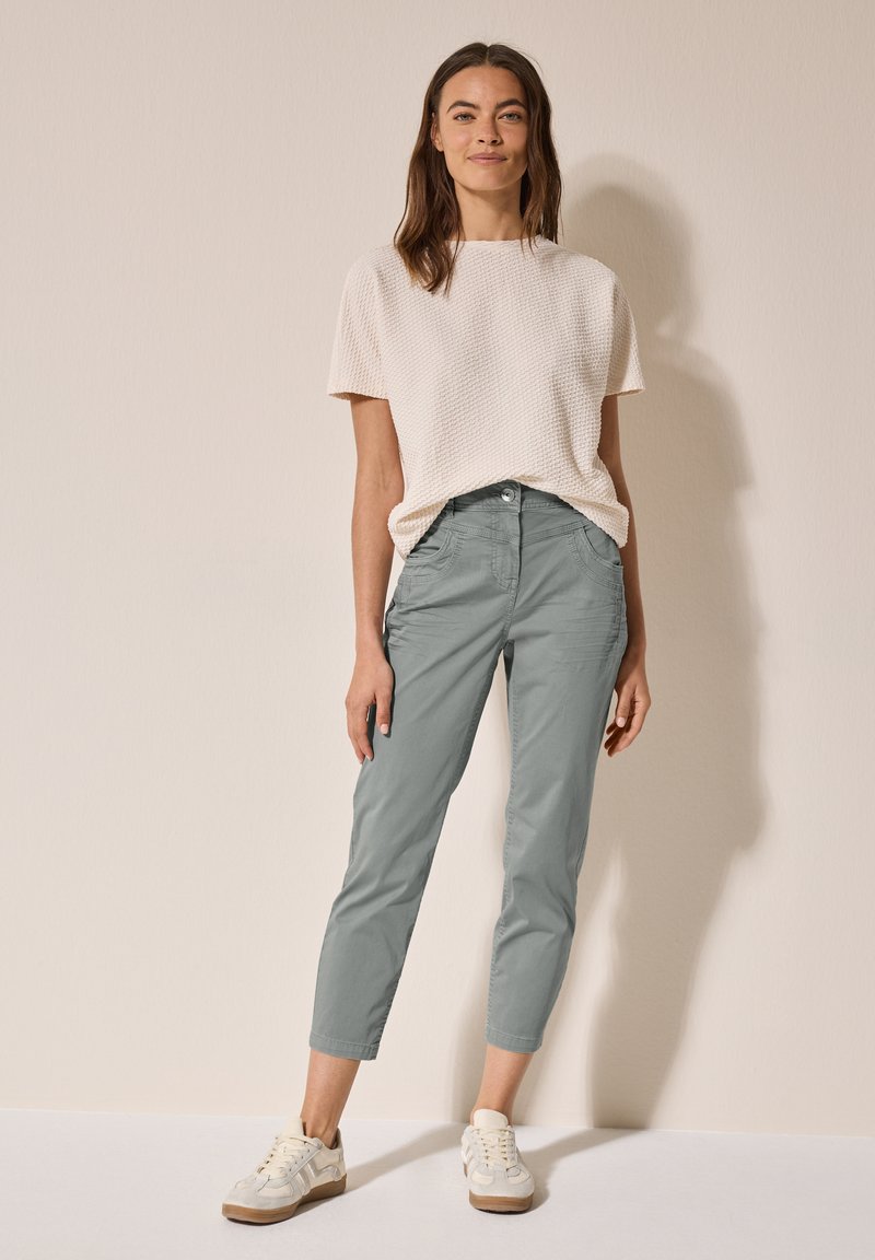 Top in maglia a trama fine di colore crema chiaro, abbinato a pantaloni grigi chiari a sigaretta e sneakers beige. I pantaloni presentano tasche anteriori e una chiusura a bottone.