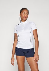 Polo bianco con accenti neri sui lati, maniche corte e pattina dei bottoni, abbinato a pantaloni corti blu navy. Texture liscia, vestibilità atletica.