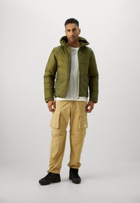 Doudoune vert olive avec capuche, fermeture éclair à l'avant et poches latérales, associée à un pantalon cargo beige clair et des bottes de randonnée noires.