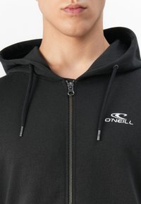 Hombre que lleva una sudadera con cremallera negra, con capucha, cordones y el logo blanco de O'Neill en el pecho.