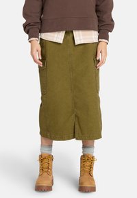 Falda midi verde oliva de mezclilla con dos bolsillos laterales, combinada con un sudadera marrón y botas de encaje color marrón claro. Tejido texturizado.