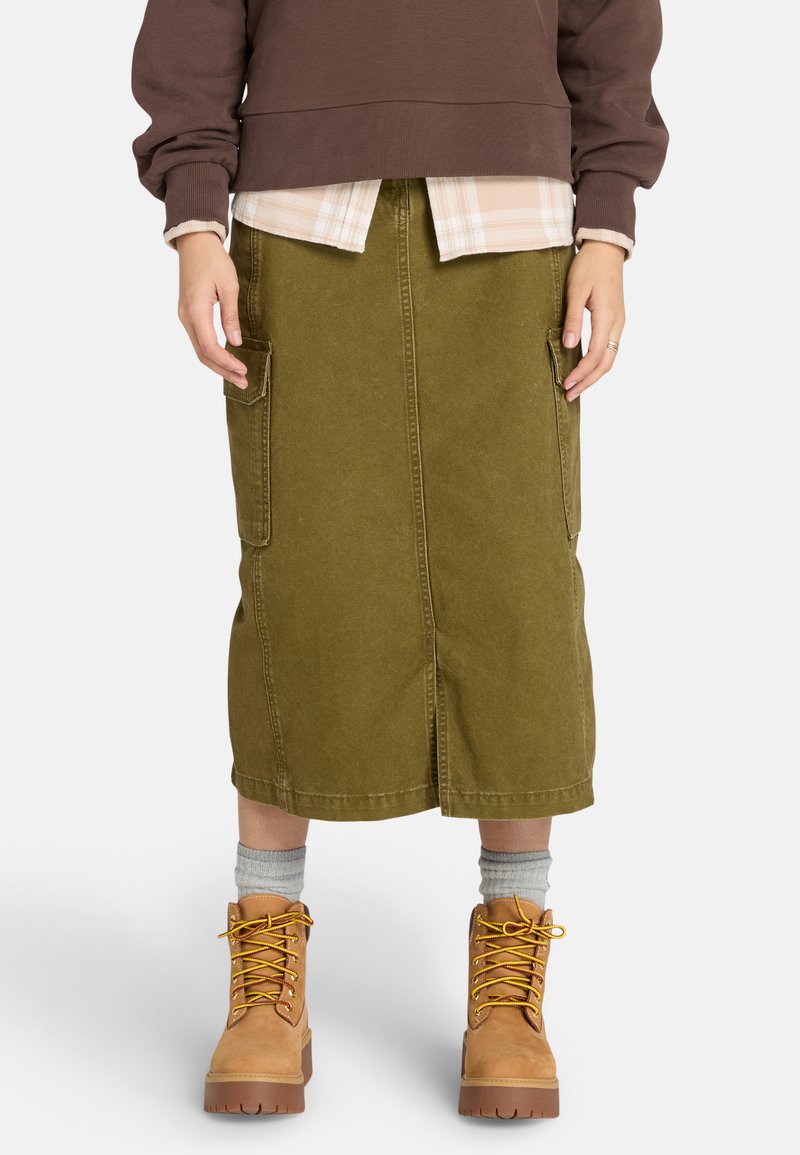 Falda midi verde oliva de mezclilla con dos bolsillos laterales, combinada con un sudadera marrón y botas de encaje color marrón claro. Tejido texturizado.