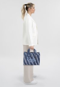 Blue tie-dye tote bag with embroidered "Coach New York" text, canvas material, short handles, and a tag. Worn with a white blazer and beige pants.