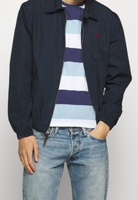 Giacca leggera blu navy con polsini elasticizzati, chiusura con zip e tasche laterali, indossata sopra una t-shirt a righe blu, bianca e viola, abbinata a jeans.