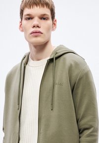 Felpa con cappuccio verde oliva con chiusura lampo e coulisse, presenta un marchio discreto "BOSS", indossata sopra un maglione a coste color crema. Texture liscia.