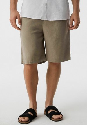 Mężczyzna noszący beżowe krótkie spodenki do kolan, białą koszulę zapinaną na guziki oraz czarne sandały, stojący na jednolitym tle.