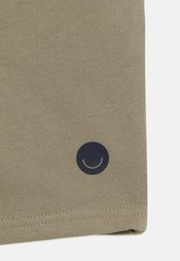 Khaki kokvilnas audums ar gludu tekstūru, kurā ir tumši zils apļa logo ar smaidīgu seju, novietots apakšējā stūrī.