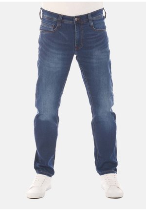 Persona con jeans blu dritti e sneakers bianche, in piedi su uno sfondo bianco uniforme. La parte superiore del corpo non è visibile.