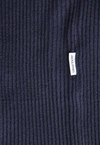 Tamsiai mėlynos tekstūruotos mezgimo medžiagos audinys; ant šono yra mažas baltas etiketė su užrašu "JACK & JONES".