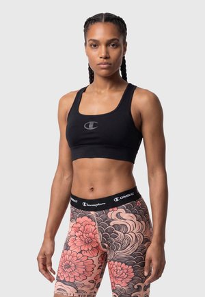 Femme en forme avec des cheveux tressés porte un soutien-gorge de sport noir et un short taille haute à motifs floraux et ondulés, debout devant un fond uni.