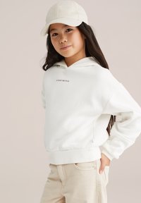 Felpa bianca con tasca frontale, con la scritta "SUNDAY BRUNCH". Indossata con pantaloni beige chiari e un cappellino di colore chiaro. Texture liscia.