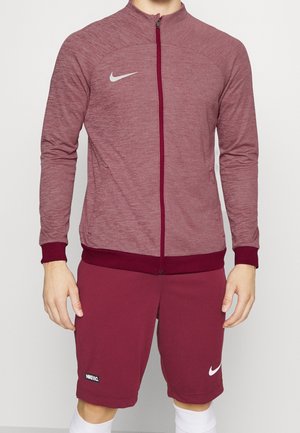 Homme portant une veste zippée Nike bordeaux et un short assorti avec des logos Nike blancs, debout devant un fond uni.