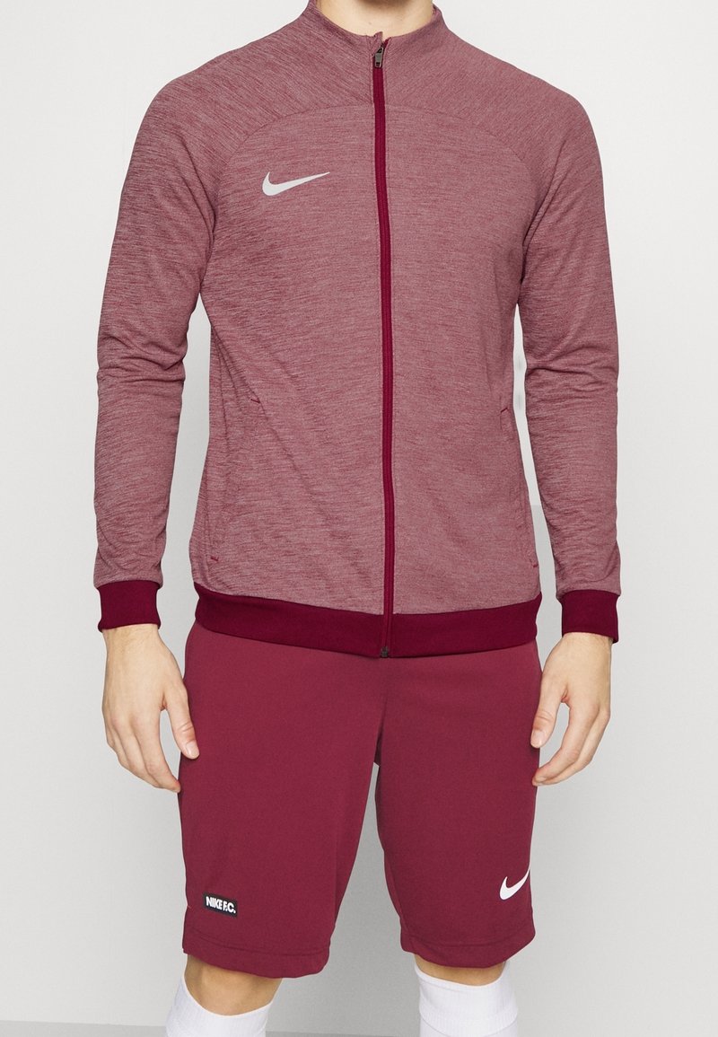 Homme portant une veste zippée Nike bordeaux et un short assorti avec des logos Nike blancs, debout devant un fond uni.